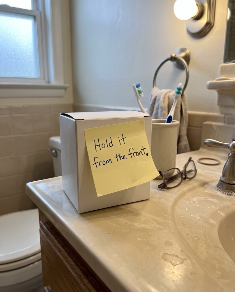 A small box on a bathroom counter with a yellow sticky note: Hold it from the front.